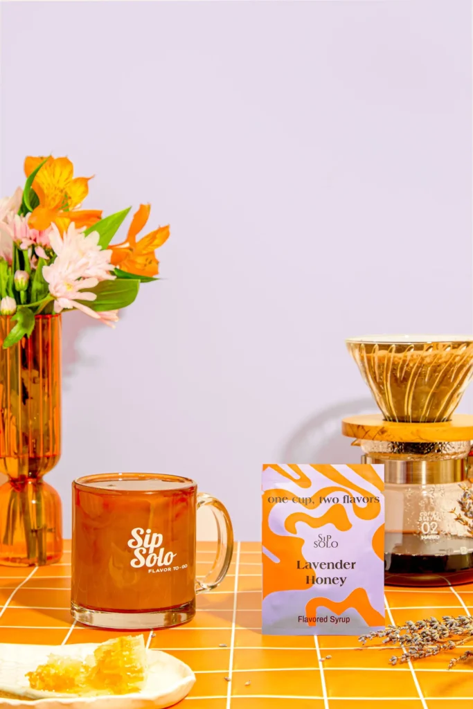 An amber mug with the words Sip Solo written on it in white lettering next to a vase of flowers, and a packet of Sip Solo Lavender Honey.