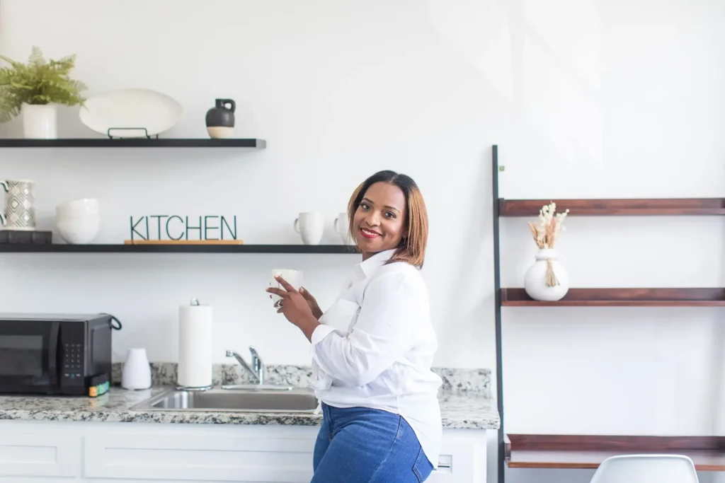 A photo of Tammy Bowser, wearing a white button-down shirt and blue jeans, standing in a kitchen holding a white mug.